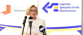 Wiceminister Machałek w białym ubraniu stoi i mówi do trzech mikrofonów na stojakach, za nią biała ściana z napisem Legnicka Specjalna Strefa Ekonomiczna.