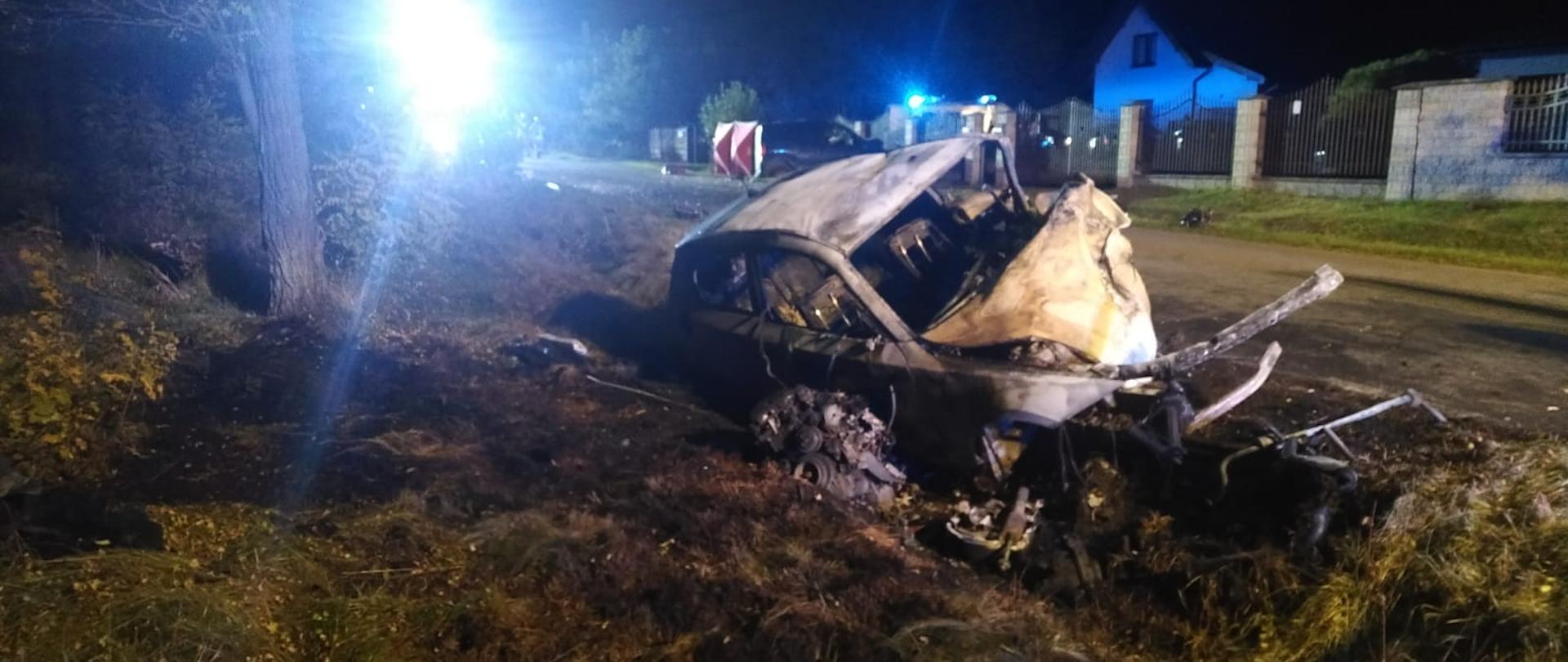 Na zdjęciu widać pojazd marki BMW. Pojazd w kolorze białym, częściowo spalony, znajdujący się w przydrożnym rowie. Za pojazdem znajduje się droga publiczna. W oddali widać zabudowania mieszkalne i drzewa. Zdjęcie wykonane w porze nocnej.