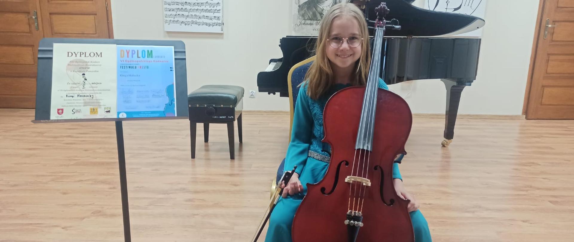 Na zdjęciu siedzi dziewczynka trzymając wiolonczelę obok ustawiono pulpit z Dyplomami w tle z napisami Dyplom Laureata i Dyplom za zajęcie II miejsca.. W tle fortepian i portrety Jana Sebastiana Bacha, oraz Logo Szkoły i partytura J. S. Bacha.
