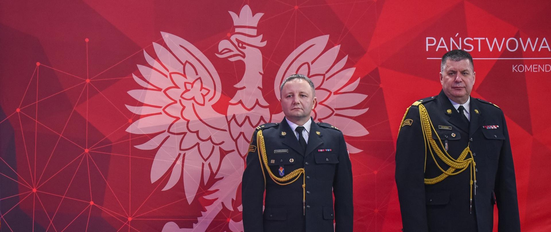 Zdjęcie przedstawia dwóch oficerów w ciemnogranatowych mundurach wyjściowych ze złotymi sznurami galowymi, stojących na baczność przodem do obiektywu. Mężczyźni znajdują się na tle dużej, czerwonej ścianki z białym godłem Polski