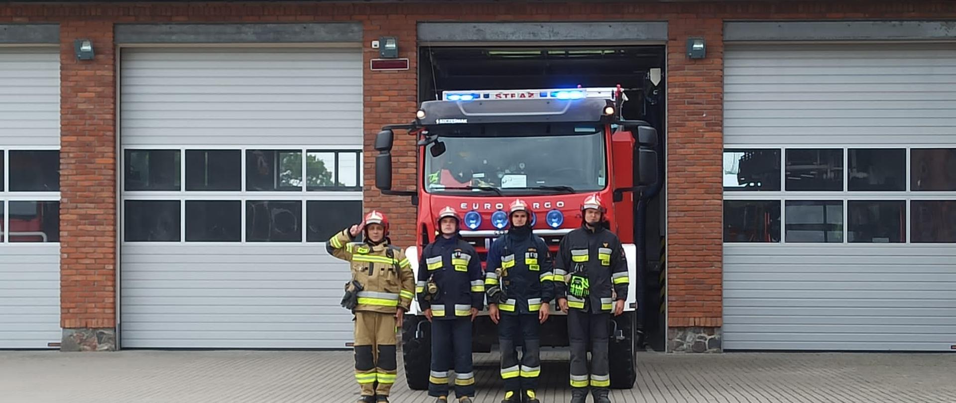 Zdjęcie przedstawia czterech strażaków Komendy Powiatowej Państwowej Straży Pożarnej w Gołdapi, z których jeden oddaje honor podczas upamiętnienia 77. rocznicę wybuchu Powstania Warszawskiego na tle pojazdu pożarniczego z włączonymi sygnałami świetlnymi i dźwiękowymi i strażnicy