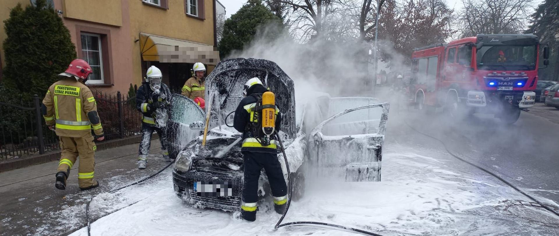 pożar samochodu na ulicy wokół dym samochód pokryty biała piana gaśniczą podniesiona maska przednia samochodu za pojazdem budynek mieszkalno usługowy koloru żółtego z tyłu samochód strażacki