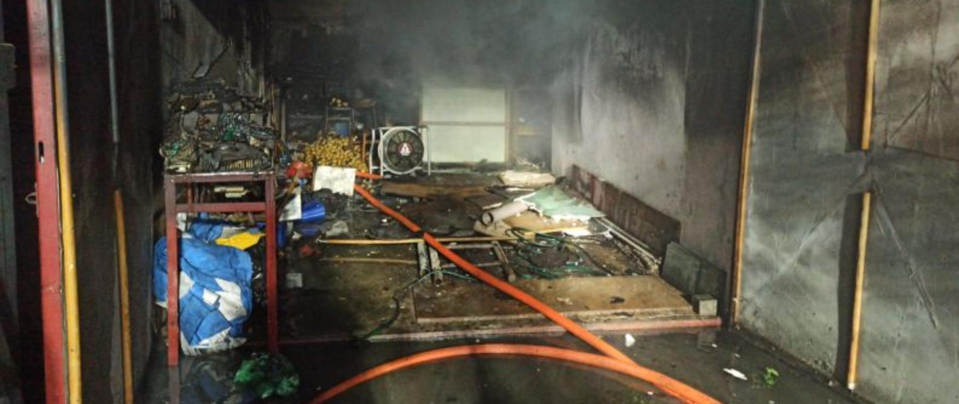 Zdjęcie wykonane w nocy, przed otwartą bramą garażu. Widać wnętrze garażu po pożarze. Pomieszczenie ma okopcone ściany, na środku stoi agregat oddymiający. Na podłodze leżą rozwinięte węże strażackie