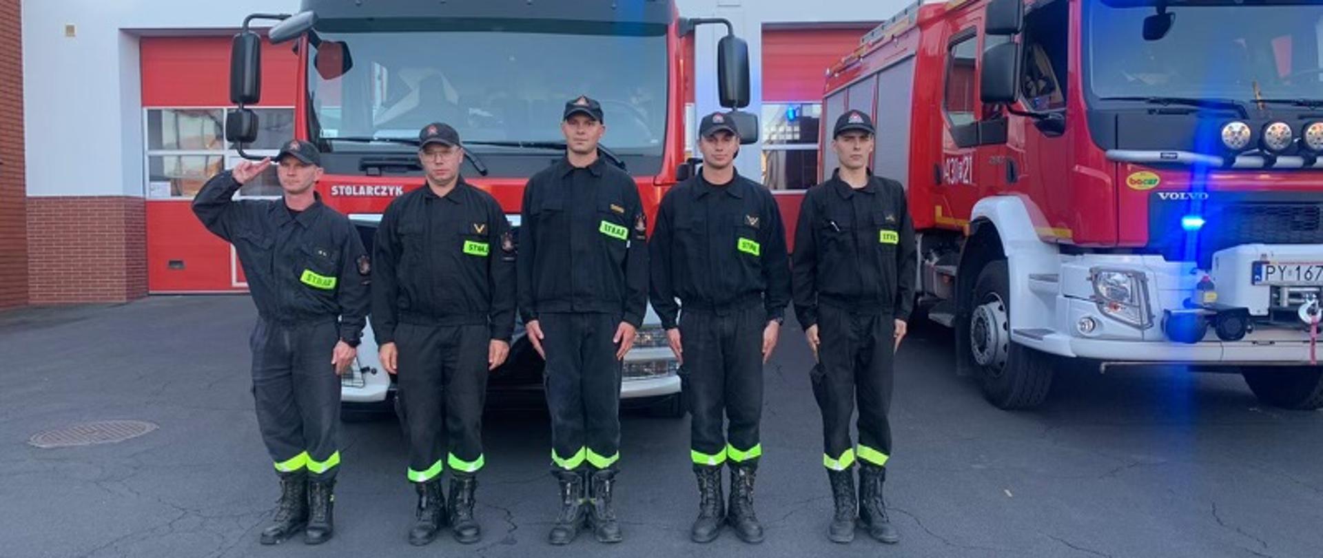 5 strażaków stoi w szeregu w pozycji na baczność. Jeden z nich oddaje honor poprzez salutowanie. W tle dwa wozy strażackie z włączonymi sygnałami świetlnymi.