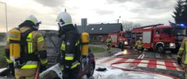 Zdjęcie na drodze publicznej. Widać auto, które uległo spaleniu. Wokół auta umundurowani ratownicy. Na drodze pozostałości piany gaśniczej. Zdjęcie wykonane w ciągu dnia.