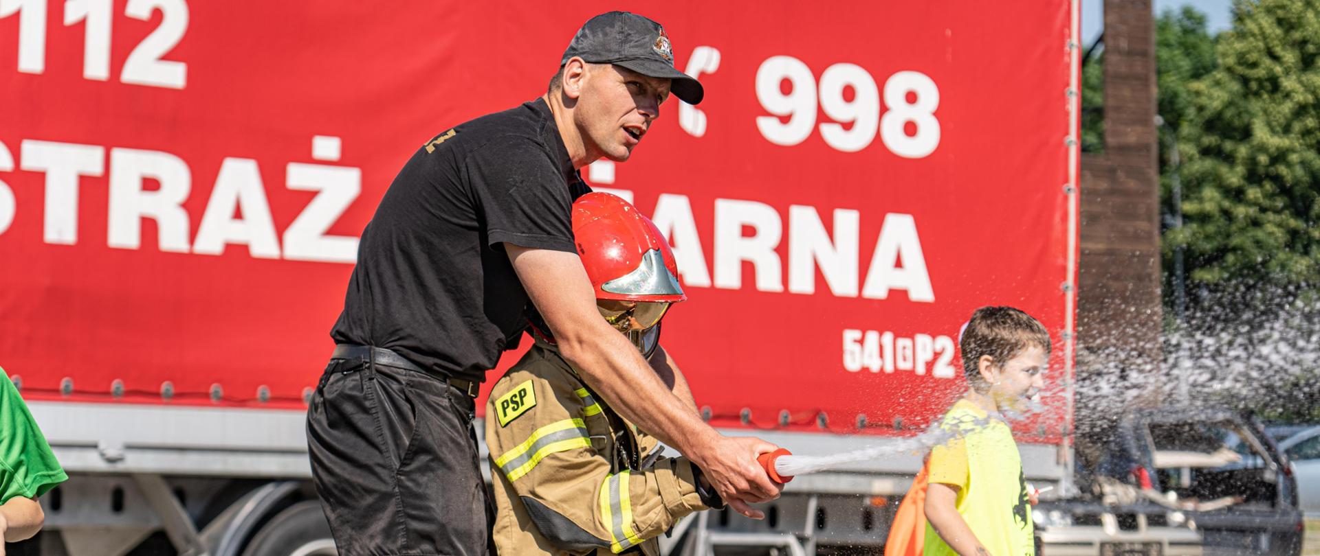 Strażak wspólnie z dzieckiem przebranym w mundur i hełm wspólnie trzymają prądownicę, z której leci woda. W tle przyczepa z napisem straż pożarna i inne dzieci.