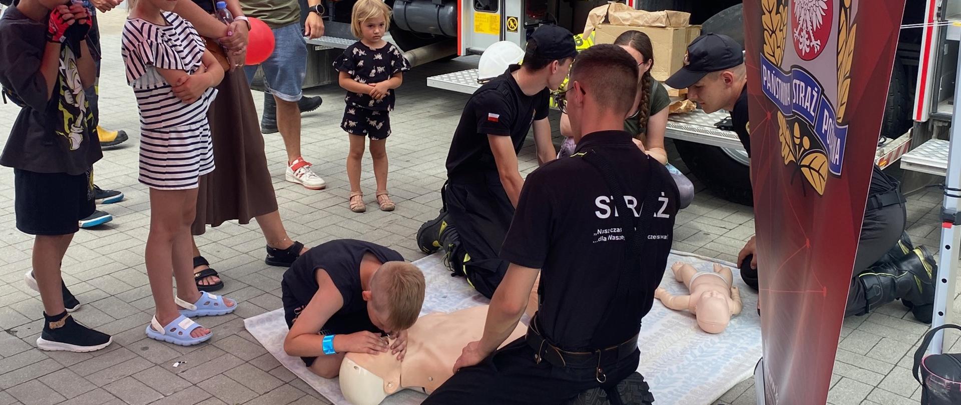 Strażacy ubrani w mundury koszarowe na kocu prezentują zasady udzielania pierwszej pomocy, obok zebrana widownia, na drugim planie samochód ratowniczo – gaśniczy.