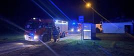 Zdjęcie przedstawia wozy strażackie z włączonymi światłami błyskowymi koloru niebieskiego stojące na drodze. Wokół stoją strażacy wykonujący zadania służbowe. 