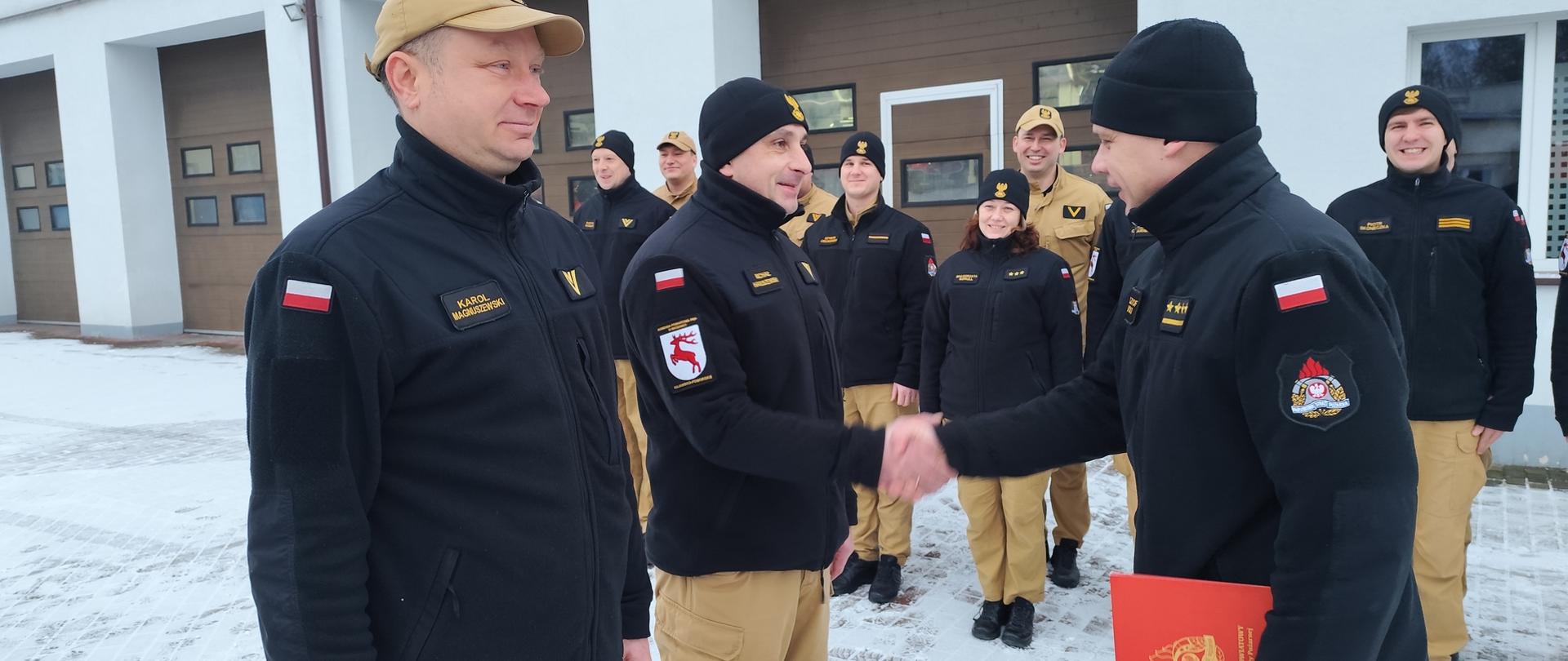 Zdjęcie przedstawia starszego kapitana Krzysztofa Natuckiego Komendanta Powiatowego PSP w Brodnicy wręczającego list gratulacyjne z okazji jubileuszu 25 lat służby i pracy starszemu ogniomistrzowi Michałowi Hanuszewskiemu. Obok niego stoi drugi jubilat PSP. Za nimi funkcjonariusze Komendy w Brodnicy.