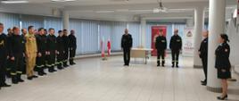 Kilkudziesięciu mężczyzn i kobiet ubranych w czarne ubrania wyjściowe, żółte sznury galowe, białe koszule, czarnym butami, oraz ubranych w czarne i żółte ubrania służbowe stoją. W tle na ścianie sztandary w gablotach szklanych, flagi biało-czerwone, rollupy w symbolem straży pożarnej oraz żaluzje koloru szarego