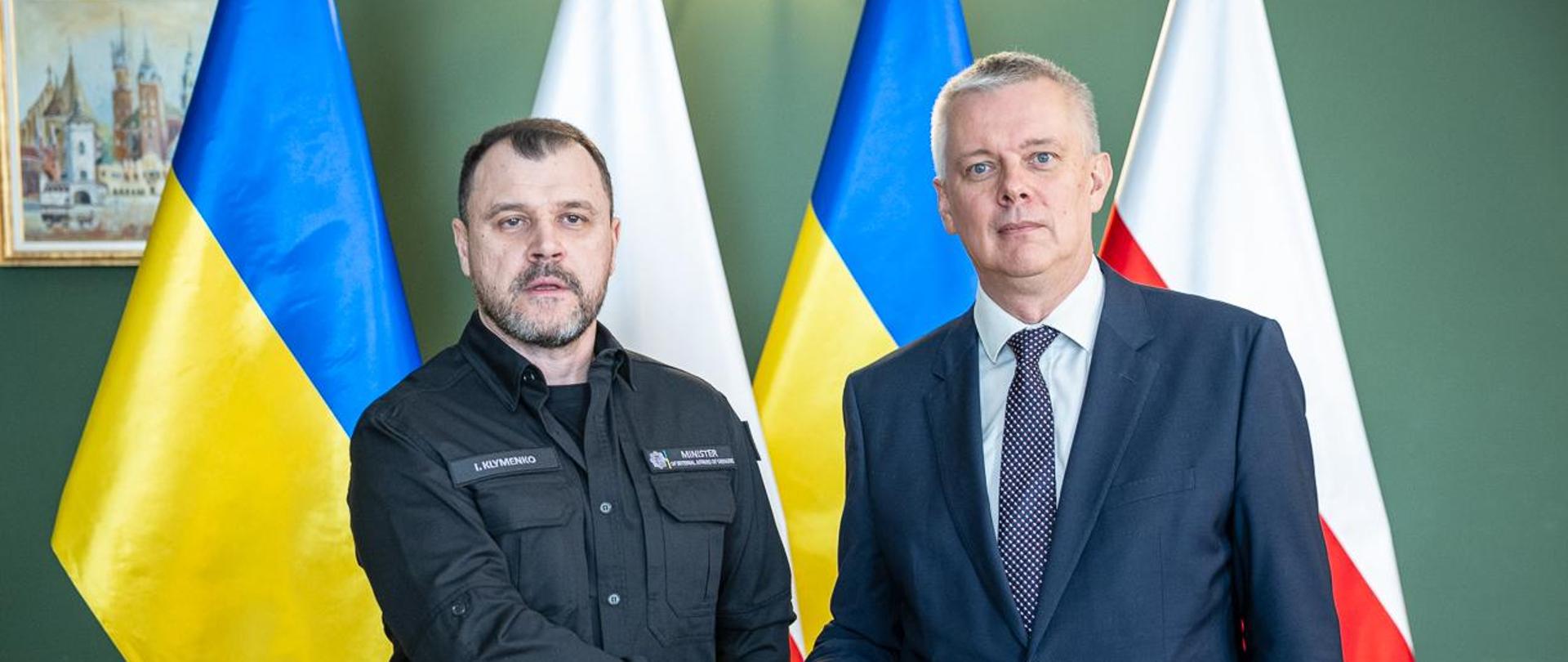 Ministrowie Tomasz Siemoniak i Ihor Kłymenko stoją na flagach polsko-ukraińskich i podają sobie ręce. 