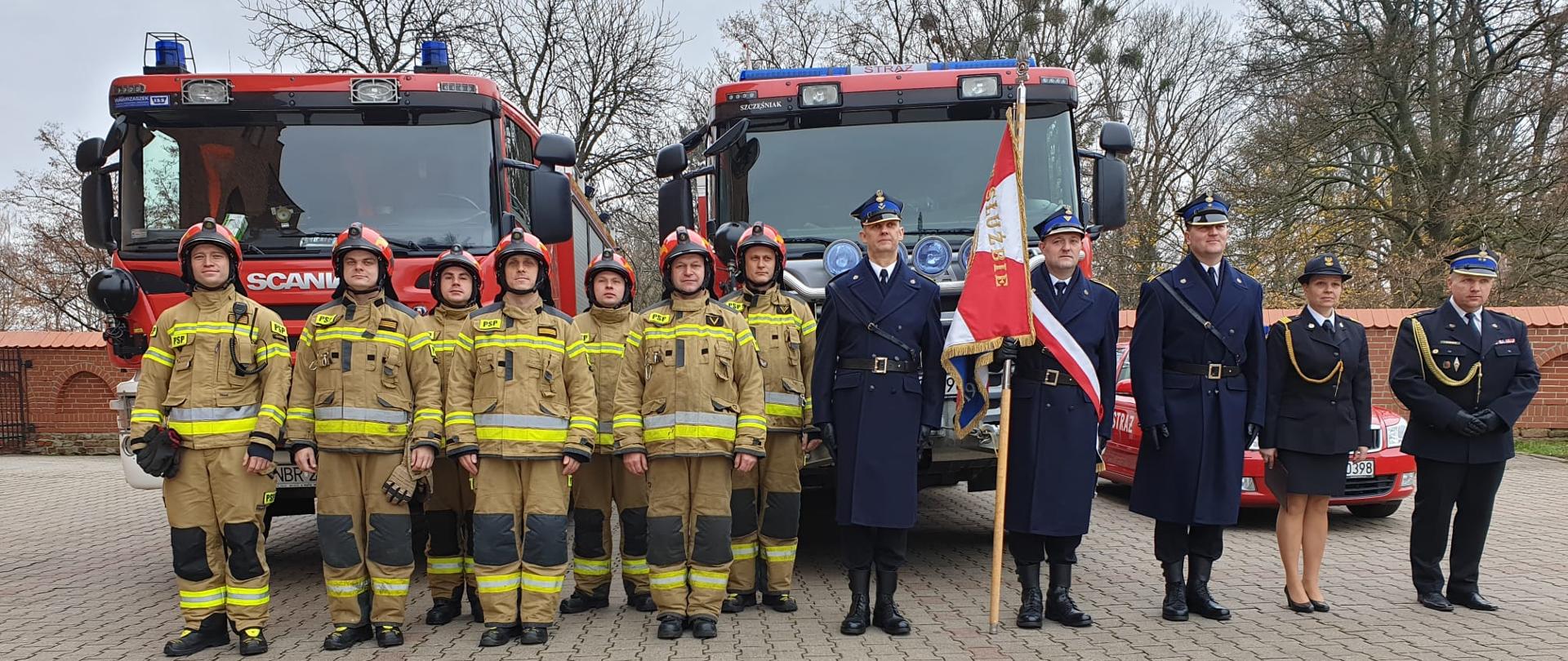 Na zdjęciu strażacy w mundurach galowych ze sztandarem oraz strażacy w mundurach bojowych w czerwonych hełmach. Z tyłu samochody strażackie.