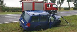 Zdjęcie przedstawia samochód osobowy z znajdujący się w przydrożnym rowie na tle pojazdu pożarniczego. Samochód uczestniczący w wypadku posiada uszkodzoną maskę oraz wystrzelone poduszki powietrzne. 