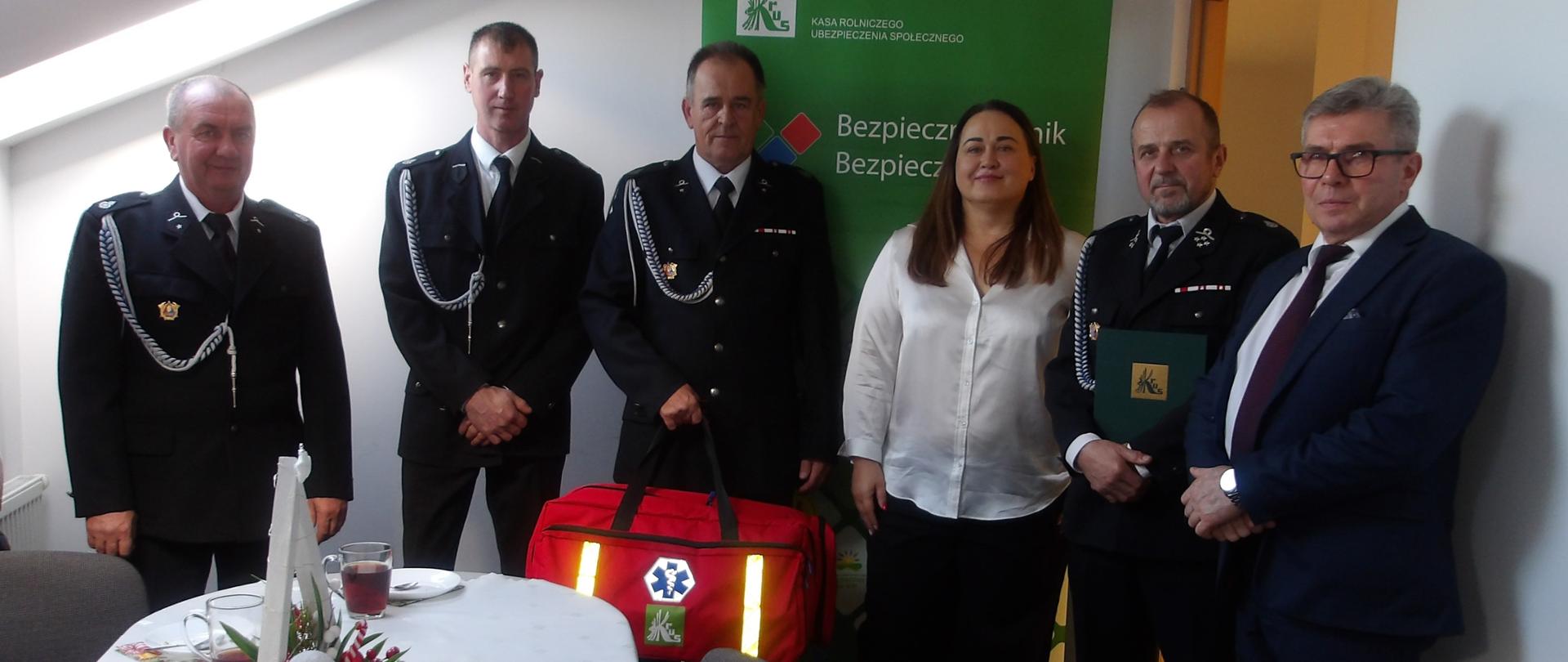 Grupa strażaków w mundurach i elegancko ubrana kobieta stoją na tle zielonego banneru z logo KRUS obok świątecznie udekorowanego stołu. Jeden osoba trzyma przekazany zestaw – czerwoną torbę R1. Po prawej stronie mężczyzna w garniturze.