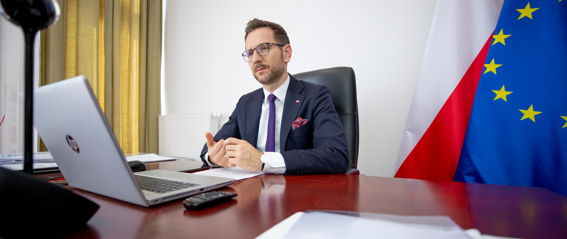 Wiceminister Waldemar Buda siedzi przy biurku przed laptopem. Za nim przy ścianie flagi PL i UE.