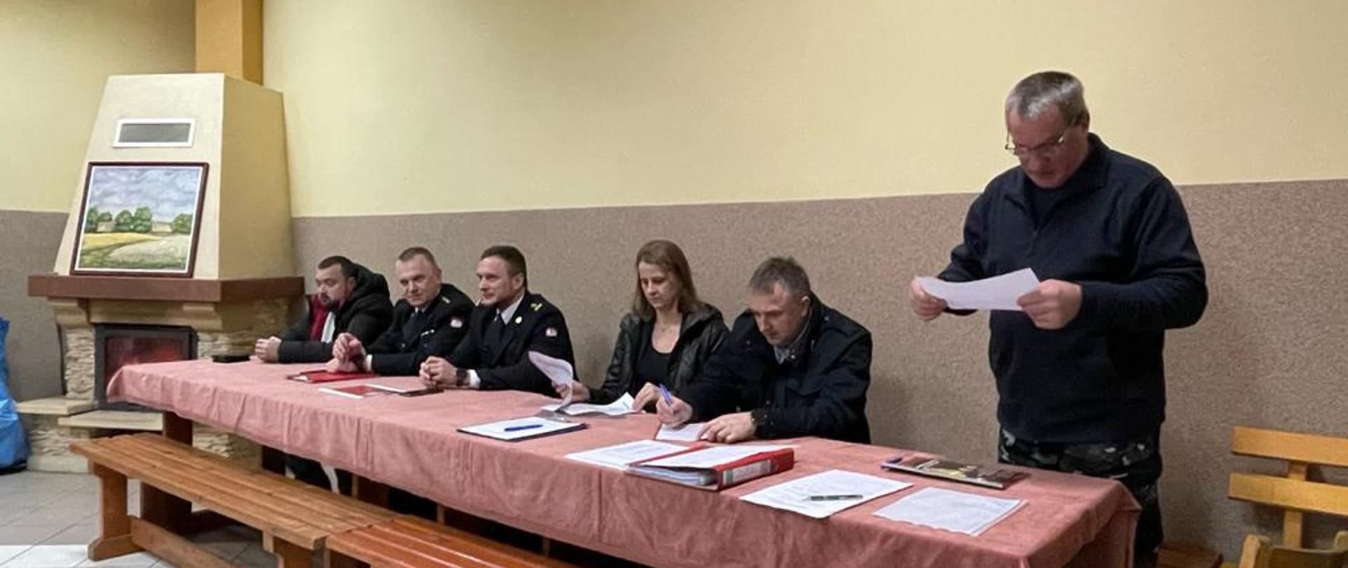 W pomieszczeniu przy stole siedzi kilka osób. Dwie z nich ubrane są w ciemne mundury.