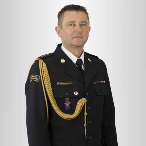 Na zdjęciu stoi mężczyzna w wieku około pięćdziesięciu lat, sfotografowany od kolan w górę, na jasnoszarym tle. Jest zwrócony lekko bokiem, ale patrzy prosto w obiektyw. Ma krótkie, ciemne włosy z lekką siwizną po bokach i spokojny, neutralny wyraz twarzy.
Ubrany jest w ciemny, elegancki mundur galowy Państwowej Straży Pożarnej. Mundur jest starannie zapięty, z białą koszulą i czarnym krawatem pod spodem. Na ramionach i kołnierzu znajdują się złote oznaczenia stopnia. Przez prawe ramię biegnie gruby, ozdobny złoty sznur, który opada na przód munduru. Na lewej piersi widoczne są drobne kolorowe wstążki odznaczeń i przypinki.
Na prawym rękawie znajduje się ciemna naszywka z napisem „Państwowa Straż Pożarna”. Całość wygląda bardzo oficjalnie — jak portret służbowy wysokiego rangą strażaka lub dowódcy