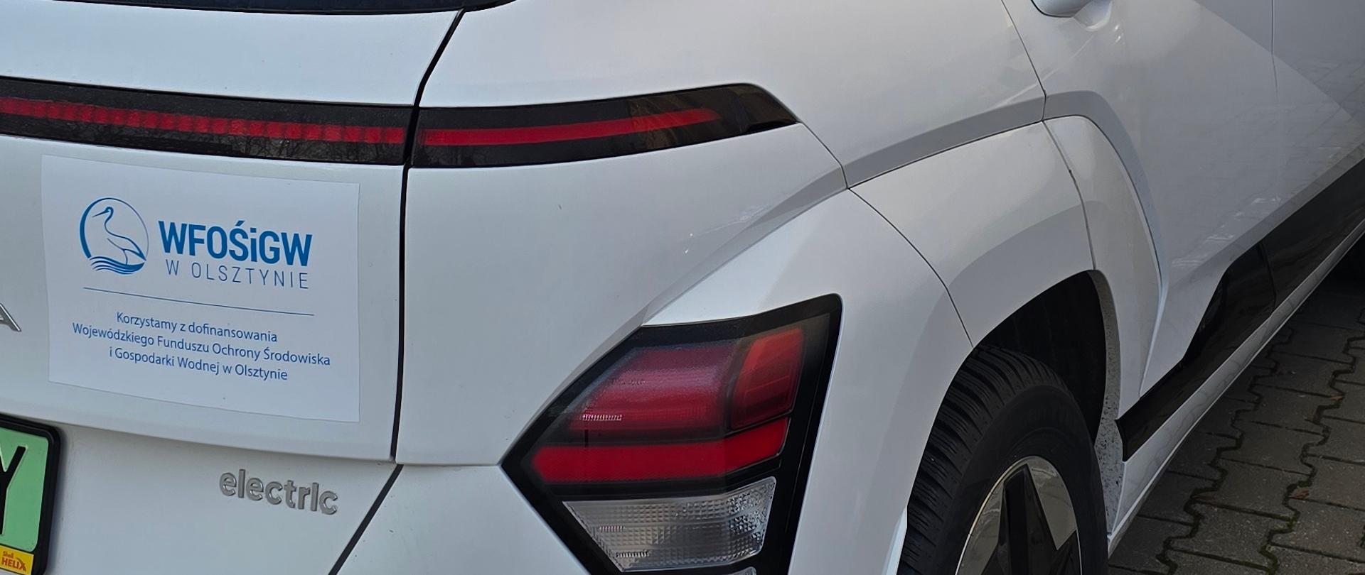Na zdjęciu widoczny jest biały samochód elektryczny zaparkowany na kostce brukowej, obok jasnej ściany budynku. Na tylnej części pojazdu znajduje się naklejka z logo WFOŚiGW w Olsztynie i informacją o dofinansowaniu. W tle widoczne są trzy maszty z flagami w kolorze białym i pomarańczowym oraz fragmenty zabudowy i drzewa. Niebo jest jasne, bezchmurne, co sugeruje słoneczną pogodę.