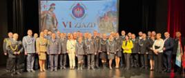Zdjęcie przedstawia pamiątkową fotografię grupową wykonaną na scenie podczas VI Zjazdu Sprawozdawczo-Wyborczego Oddziału Powiatowego ZOSP RP w Braniewie, który odbył się 17 października 2026 roku w Braniewskim Centrum Kultury.
W tle, na dużym ekranie multimedialnym, widnieje oficjalny plakat zjazdu z napisem „VI ZJAZD ODDZIAŁU POWIATOWEGO ZOSP RP W BRANIEWIE”. Po lewej stronie grafiki umieszczono wizerunek św. Floriana – patrona strażaków, w centrum znajduje się godło Związku Ochotniczych Straży Pożarnych RP, a po prawej stronie widoczny jest nowoczesny wóz strażacki.
Na scenie stoi liczny, wieloosobowy rząd uczestników zjazdu ustawionych przodem do obiektywu.
