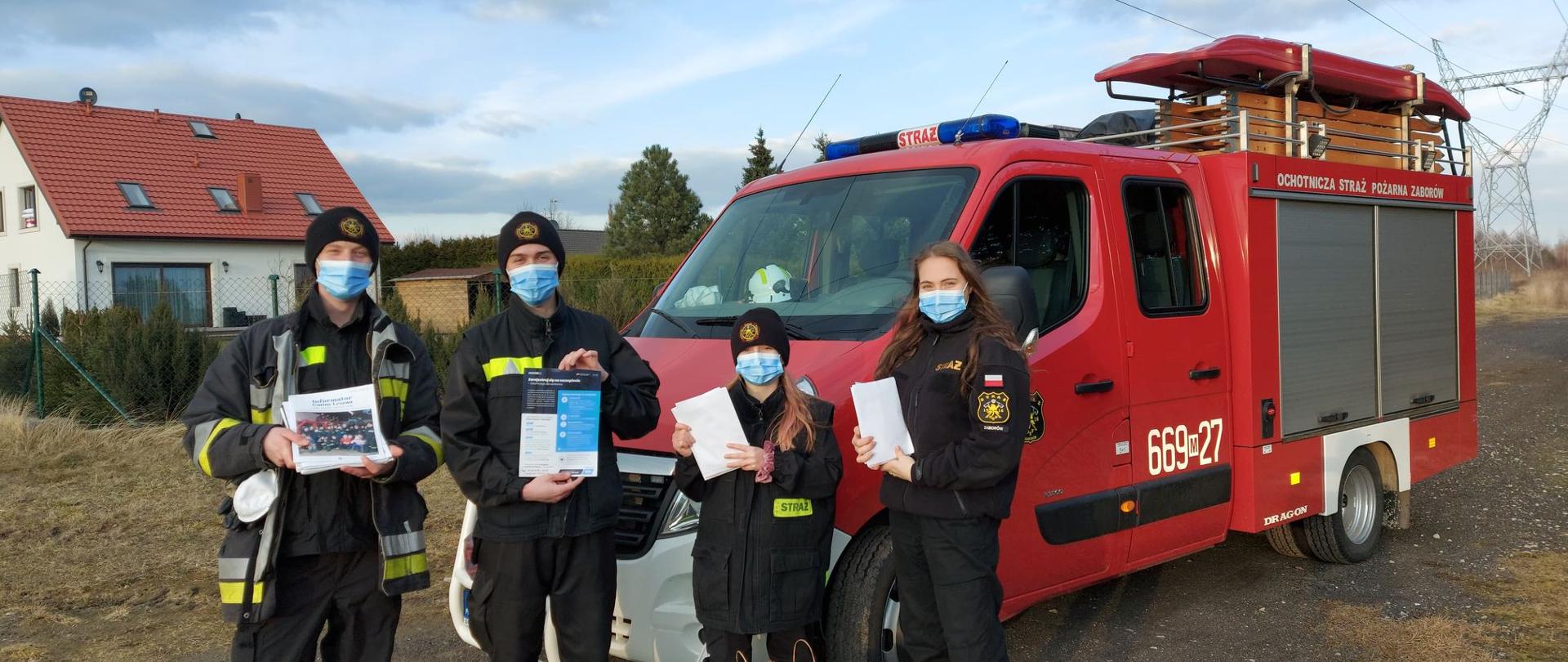 Strażacy stoją z ulotkami na tle samochodu strażackiego