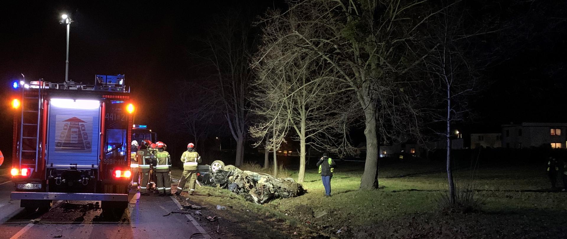 Wypadek DK 45 Gwoździce. Zdjęcie zrobione nocą przedstawia samochód pożarniczy stojący na prawym pasie drogi, oświetlający za pomocą masztu miejsce zdarzenia. Na prawo od samochodu stoją strażacy w ubraniach specjalnych i hełmach. W środku w rowie wrak samochodu po dachowaniu. Obok wraku policjant podczas wykonywania czynności służbowych. W tle pole oraz podświetlone przydrożne drzewa.