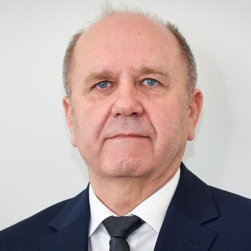 Mężczyzna w ciemnej marynarce, białej koszuli i ciemnym krawacie, z krótko przyciętymi włosami.