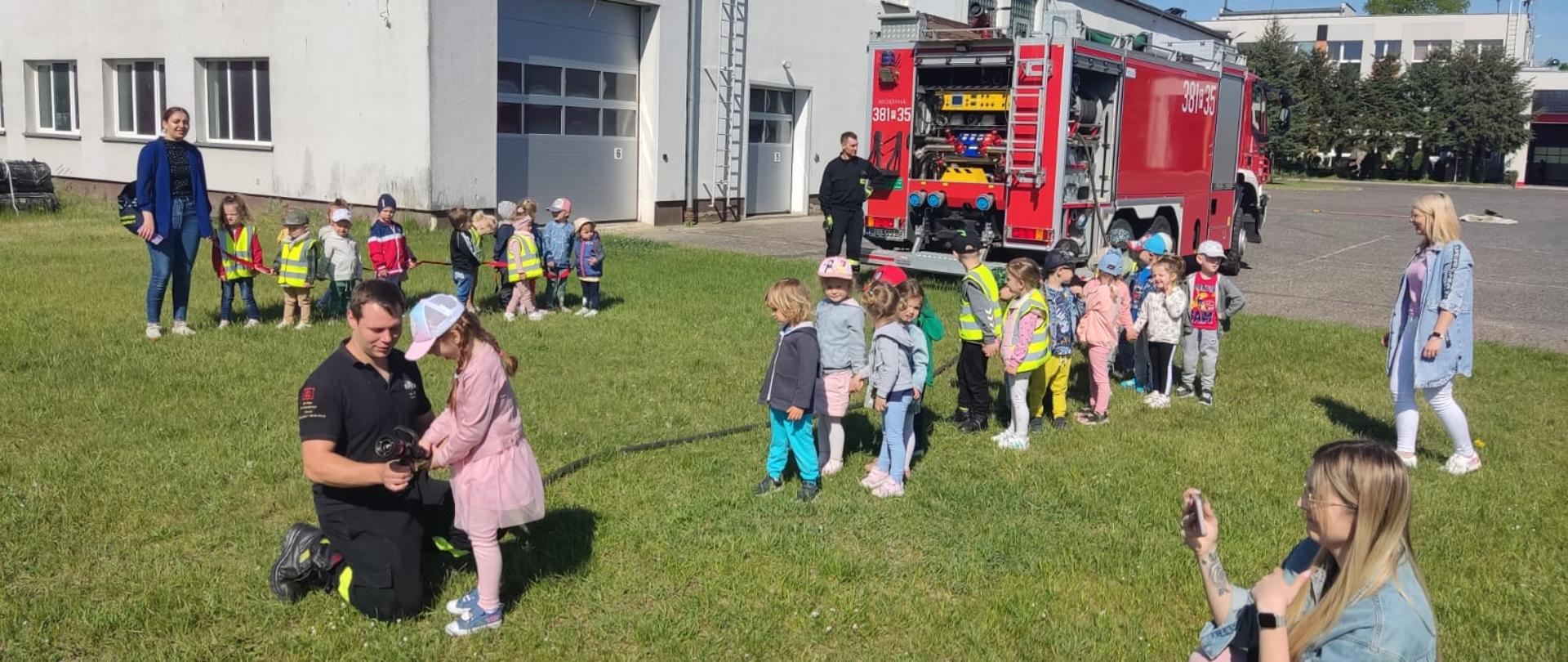 Strażacy, dzieci i osoby cywilne stoją na ziemi w tle samochód strażacki i budynki.