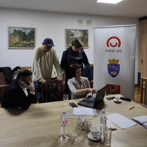 Przy stole stoją dwaj mężczyźni oraz siedzi mężczyzna i kobieta i patrzą na ekran komputera stojącego przed kobietą; obok stołu stoi baner z logo Polskiej pomocy i dwoma herbami miast