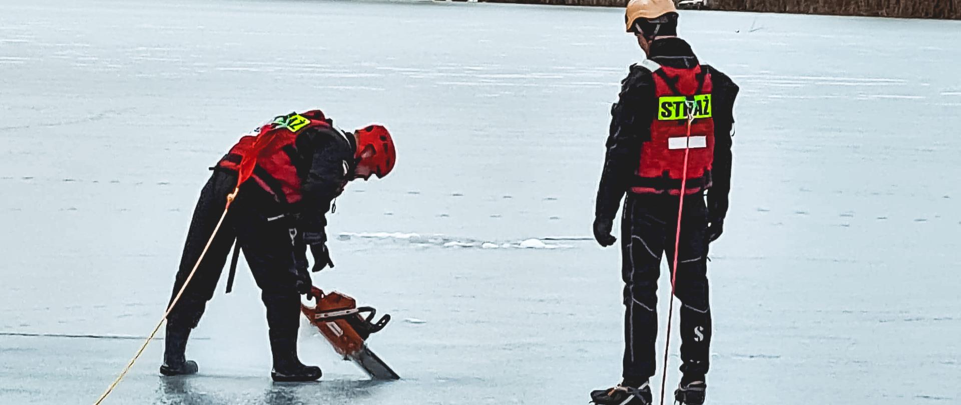 Zdjęcie przedstawia dwóch strażaków w skafandrach do pracy w wodzie wykonujących otwór w lodzie