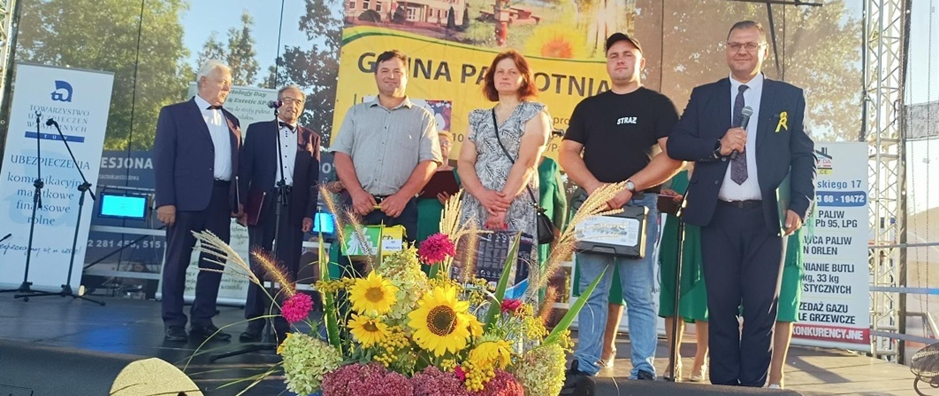 Grupa osób stojących na scenie podczas uroczystości plenerowej. W tle znajduje się baner z napisem „GMINA PAPROTNIA”, a także inne reklamy i banery sponsorów. Na środku przed sceną ustawiono białą skrzynkę z dużym, kolorowym bukietem kwiatów. Trzy osoby stojące na pierwszym planie trzymają w ręku nagrody, czwarty mężczyzna trzyma w ręku mikrofon. Za nimi na drugim planie stoi dwóch mężczyzn w garniturze i kobiety w zielonych sukienkach. Osoby stojące w pierwszym rzędzie patrzą w stronę aparatu.