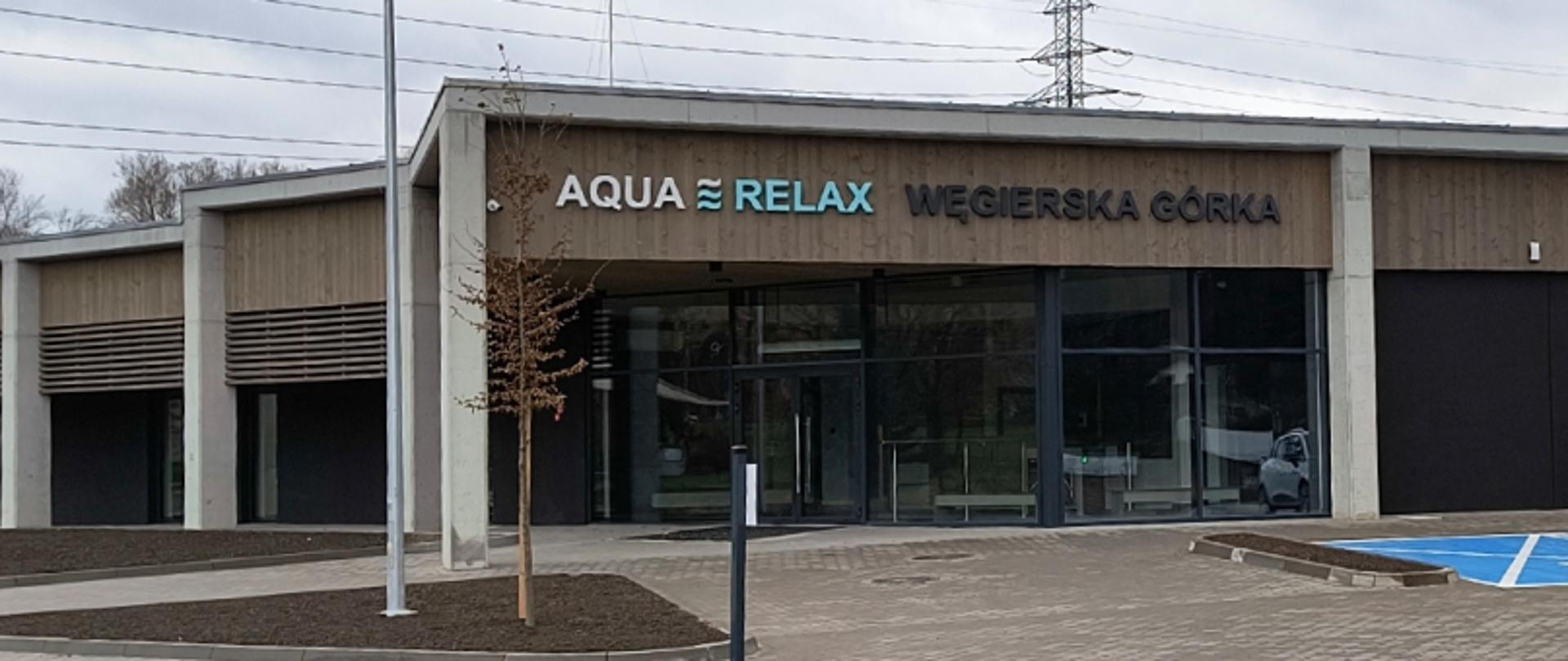 Nowoczesny budynek z napisem „AQUA RELAX WĘGIERSKA GÓRKA” nad wejściem; przeszklona fasada, drewniane panele, drzewko i oznaczone miejsce parkingowe.
