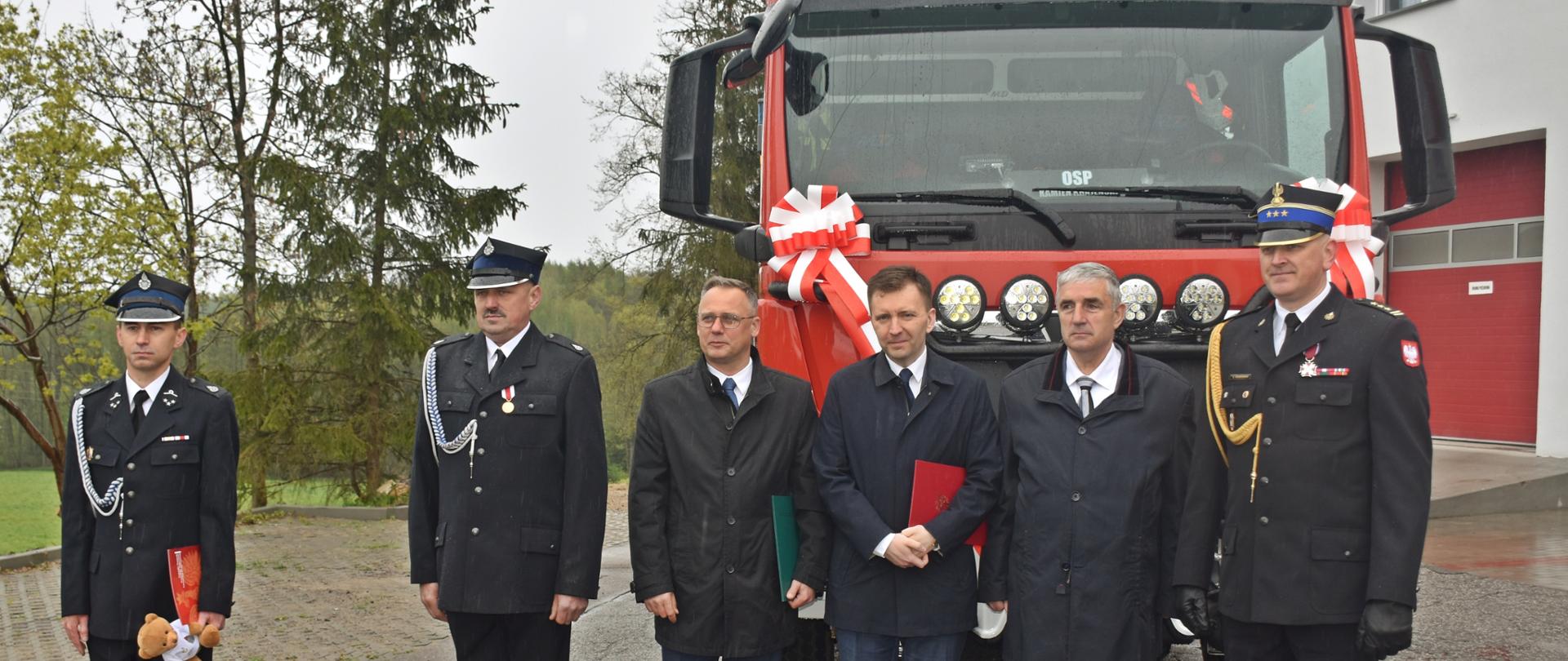 Zdjęcie wykonano w ciągu dnia podczas uroczystości gminnych obchodów dnia strażaka 2023 w Kamieniu Krajeńskim. sześcioro mężczyzn ubranych w mundury wyjściowe i garnitury stoją przed samochodem ratowniczo-gaśniczym MAN. 
