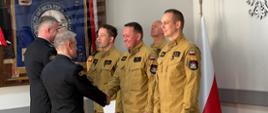 Komendant wręcza awanse i gratuluje strażakom poprzez uścisk dłoni podczas uroczystości w siedzibie Komendy Powiatowej PSP w Mławie; obok stoi poczet funkcjonariuszy oraz flaga Polski.