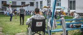 na podwórku obok ośrodka dla podopiecznych znajduje się grupa mężczyzn, którzy grają w piłkę siatkową. na wózku inwalidzkim z naklejką Polska pomoc siedzi mężczyzna