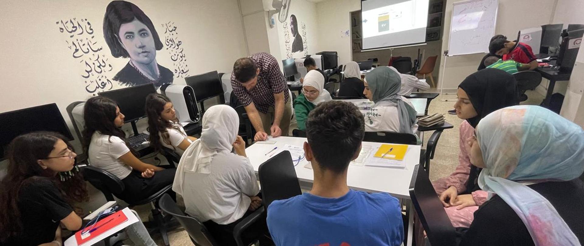 W pracowni komputerowej siedzi kilkanaście młodych kobiet i mężczyzn, jeden mężczyzna coś pokazuje zaś reszta go obserwuje