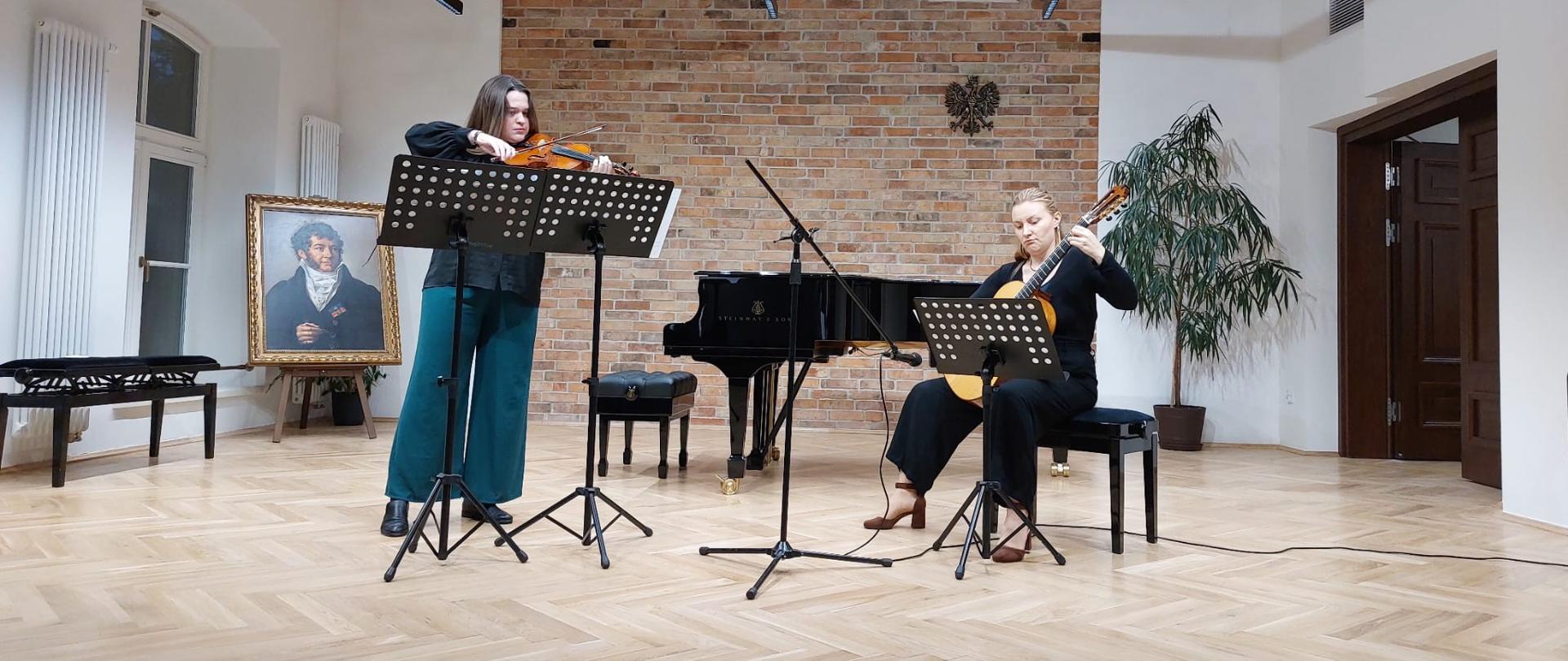 Dwie osoby wykonują duet na scenie: jedna gra na skrzypcach, druga na gitarze klasycznej; przed nimi stoją pulpity z nutami.