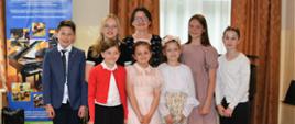 Zdjęcie uczniów klasy fortepianu po audycji pianistycznej. Stoją wraz z nauczycielką w dwóch rzędach, za nimi baner unijny, okno przysłonięte firaną i dwoma zasłonami, z prawej strony pulpit.