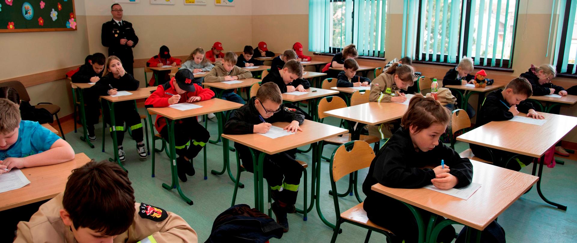 Widok sali - uczestnicy siedzą w ławkach i rozwiązują test pisemny.