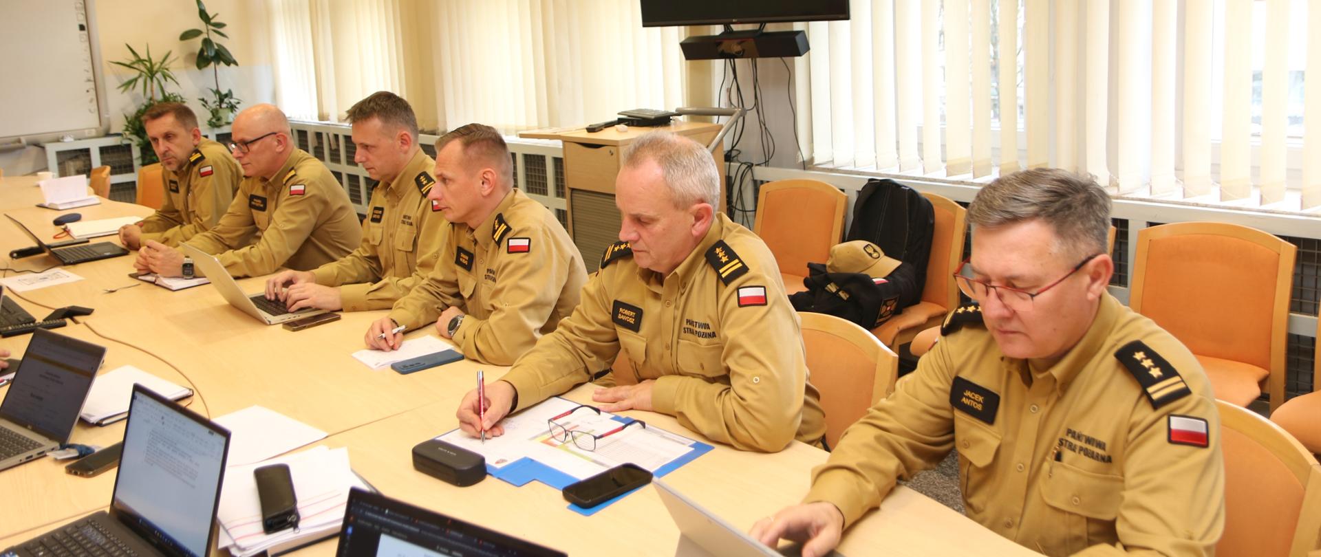 Sześciu strażaków PSP w mundurach służbowych siedzi przy długim stole konferencyjnym z laptopami i notatkami, skupieni na spotkaniu. W tle duży telewizor oraz krzesła i zasłony.