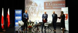 Trzej mężczyźni stoją na scenie, kobieta mówi do mikrofonu, w tle baner z napisem "105. rocznica Bitwy Niemeńskiej" i wizerunkiem Józefa Piłsudskiego siedzącego na koniu.