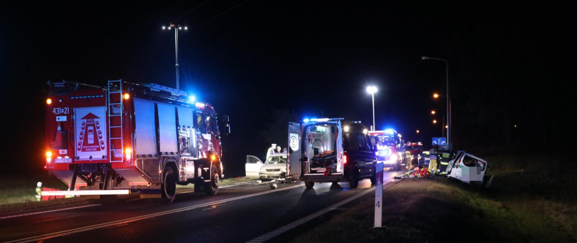Wóz strażacki oraz karetki pogotowia podczas wypadku. Wóz strażacki oświetla z masztu teren działań. Obok karetki stoi stoją samochody osobowe biorące udział w zdarzeniu. Jeden z nich znajduje się na poboczu. 