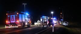 Wóz strażacki oraz karetki pogotowia podczas wypadku. Wóz strażacki oświetla z masztu teren działań. Obok karetki stoi stoją samochody osobowe biorące udział w zdarzeniu. Jeden z nich znajduje się na poboczu. 