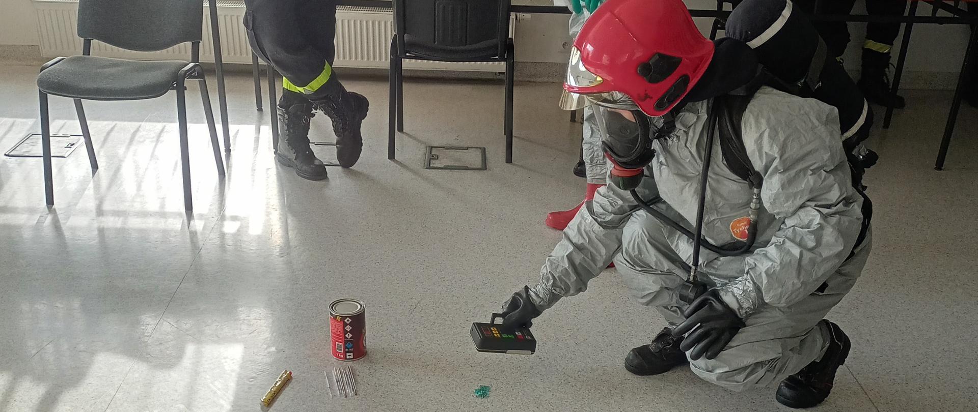 Zdjęcie przedstawia strażaka podczas szkolenia z ratownictwa chemiczno - ekologicznego. Szkolenie odbywa się na świetlicy JRG. Strażak przy pomocy urządzeń pomiarowych sprawdza substancję niebezpieczną. 