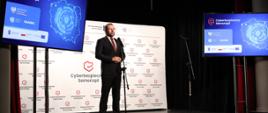 Minister funduszy i polityki regionalnej Grzegorz Puda stoi na scenie przy mikrofonie, za jego plecami baner z multiplikacją "Cyberbezpieczny Samorząd", a po lewej i prawej stronie ekrany z mapą Polski i napisem "Cyberbezpieczny Samorząd"