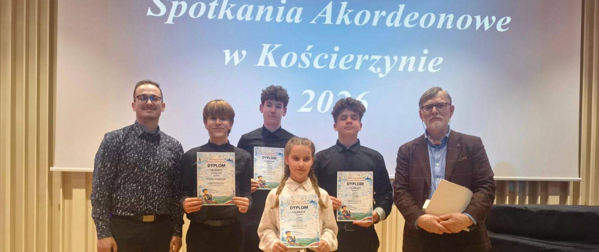 Zdjęcie pamiątkowe z XXXI Kaszubskich Spotkań Akordeonowych w Kościerzynie 2026. Na scenie stoi grupa laureatów – trzech chłopców i dziewczynka – trzymających dyplomy, w towarzystwie dwóch opiekunów lub jurorów. W tle widoczny ekran z napisem tytułowym wydarzenia.