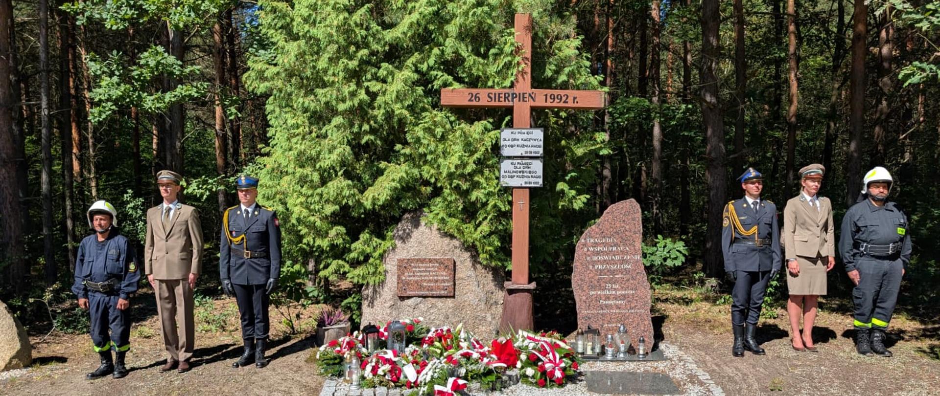 Na zdjęciu, zrobionym w słoneczny dzień, widać grupę pięciu osób w mundurach, stojących przed pomnikiem w lesie. Pomnik składa się z drewnianego krzyża z tabliczką, na której widnieje napis: "26 SIERPIEŃ 1992 r." oraz z dwóch kamiennych bloków. Pod krzyżem i kamieniami znajduje się klomb, gdzie leżą wieńce z czerwonych i białych kwiatów. Obok wieńców widać zapalone znicze. Po lewej stronie pomnika stoi trzech mężczyzn w mundurach: jeden w granatowym, drugi w zielonym, a trzeci w granatowym. Po prawej stronie stoją dwie osoby: kobieta w beżowym mundurze oraz mężczyzna w granatowym, z białym kaskiem strażackim na głowie. W tle za pomnikiem widać gęsty las.