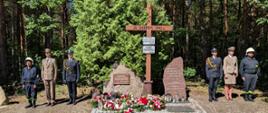 Na zdjęciu, zrobionym w słoneczny dzień, widać grupę pięciu osób w mundurach, stojących przed pomnikiem w lesie. Pomnik składa się z drewnianego krzyża z tabliczką, na której widnieje napis: "26 SIERPIEŃ 1992 r." oraz z dwóch kamiennych bloków. Pod krzyżem i kamieniami znajduje się klomb, gdzie leżą wieńce z czerwonych i białych kwiatów. Obok wieńców widać zapalone znicze. Po lewej stronie pomnika stoi trzech mężczyzn w mundurach: jeden w granatowym, drugi w zielonym, a trzeci w granatowym. Po prawej stronie stoją dwie osoby: kobieta w beżowym mundurze oraz mężczyzna w granatowym, z białym kaskiem strażackim na głowie. W tle za pomnikiem widać gęsty las.
