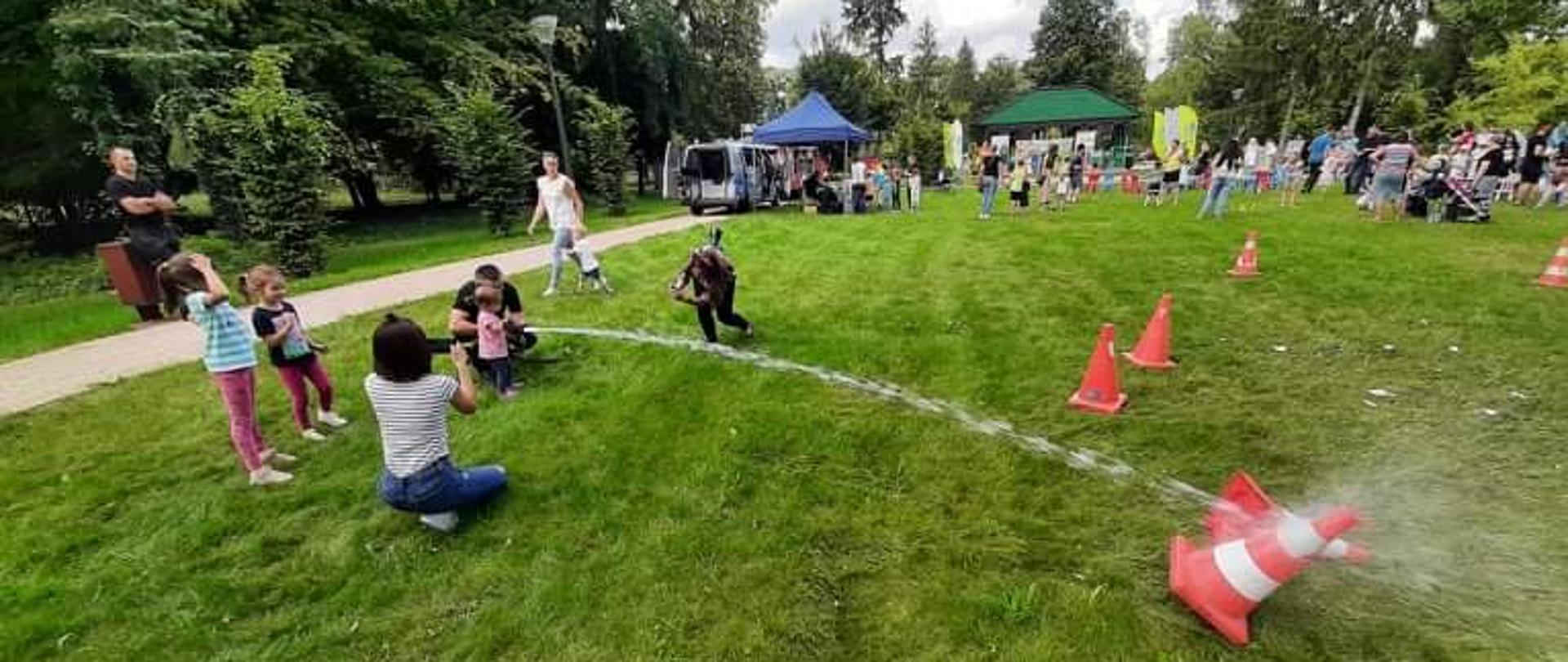 Na trawniku stoi 3 dzieci, 2 strażaków oraz 3 dorosłe kobiety. Wszyscy zaangażowani są w zabawę strącenia prądem wody pachołka stojącego w znacznej odległości od osoby podającej prąd wody na niego. Z tyłu widać stragany oraz ludzi biorących udział w pikniku. 