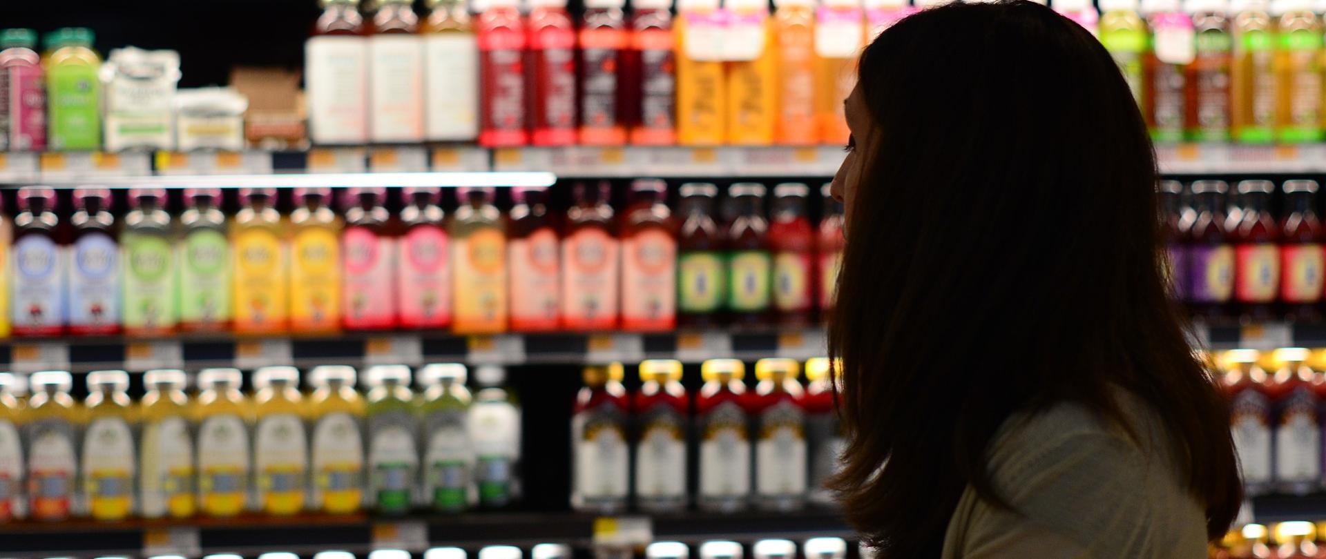 Na zdjęciu znajduje się kobieta, która robi zakupy w sklepie spożywczym. W tle są półki z napojami.