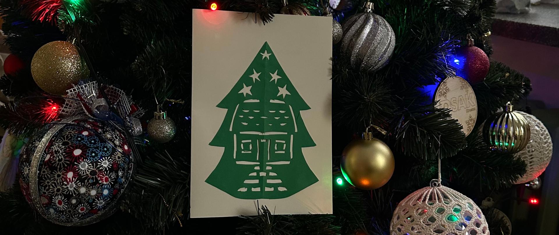 Kartka świąteczna na tle zielonej choinki z kolorowymi bombkami. 
